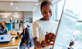A professional woman is captured engaging with a whiteboard during a business meeting in an office, accompanied by colleagues. Cooperation and productivity in a corporate atmosphere.